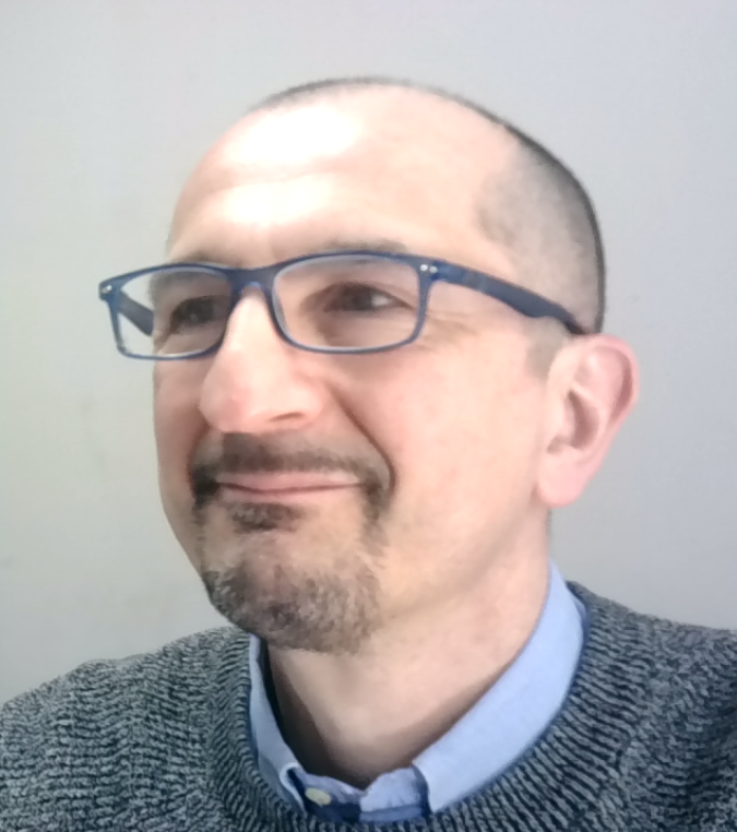 A man wearing glasses and a gray sweater smiles slightly while looking to the side against a light background.